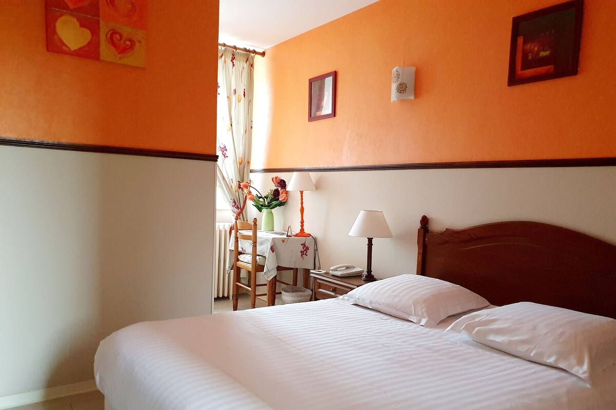 Een hotelkamer met een groot bed, een houten hoofdeinde, een tafeltje met een lamp en een stoel.