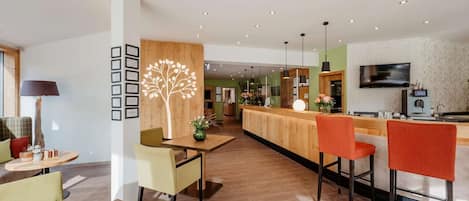 A modern hotel lobby with a wooden reception desk, seating area, and a TV mounted on the wall.