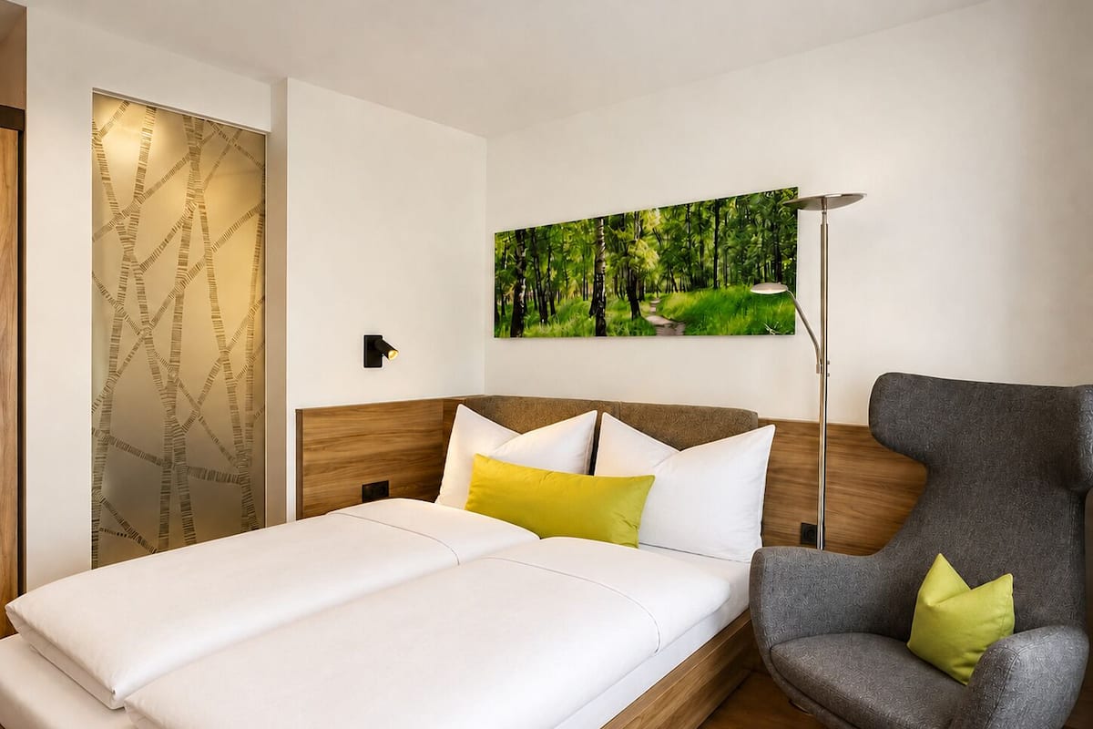 A modern hotel room with a bed, a chair, a painting, and a wall-mounted lamp.