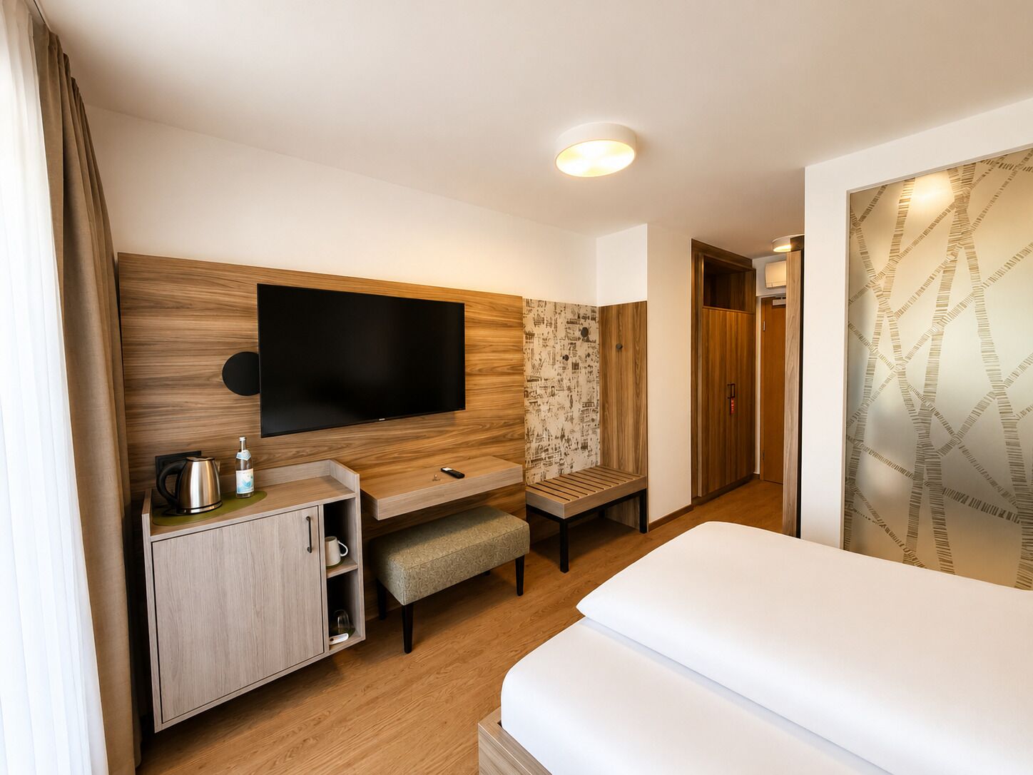 A hotel room with a bed, a TV mounted on the wall, a wooden cabinet, a bench, and a shower area.