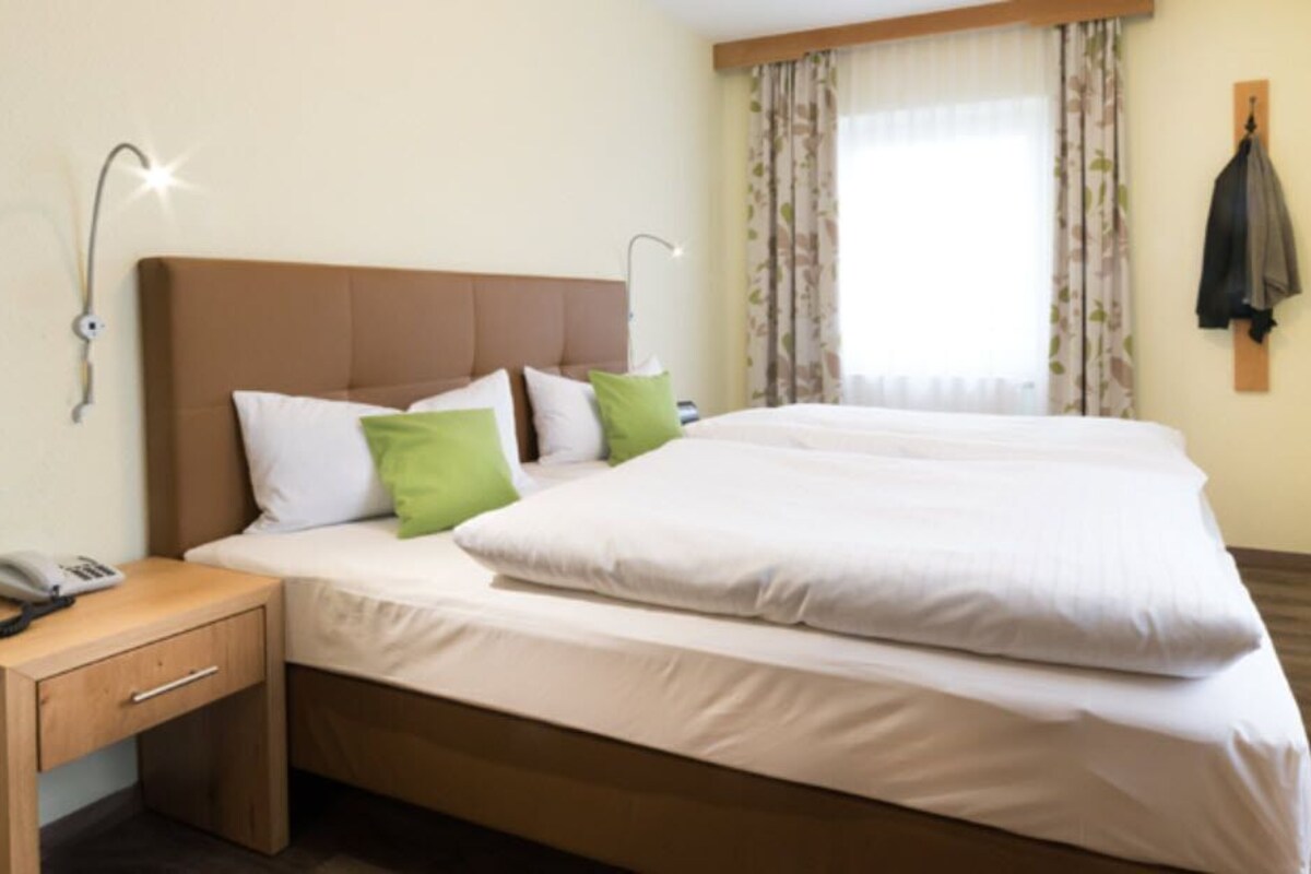 Una cama doble con ropa blanca, una mesita de noche de madera con teléfono, y un televisor montado en la pared.