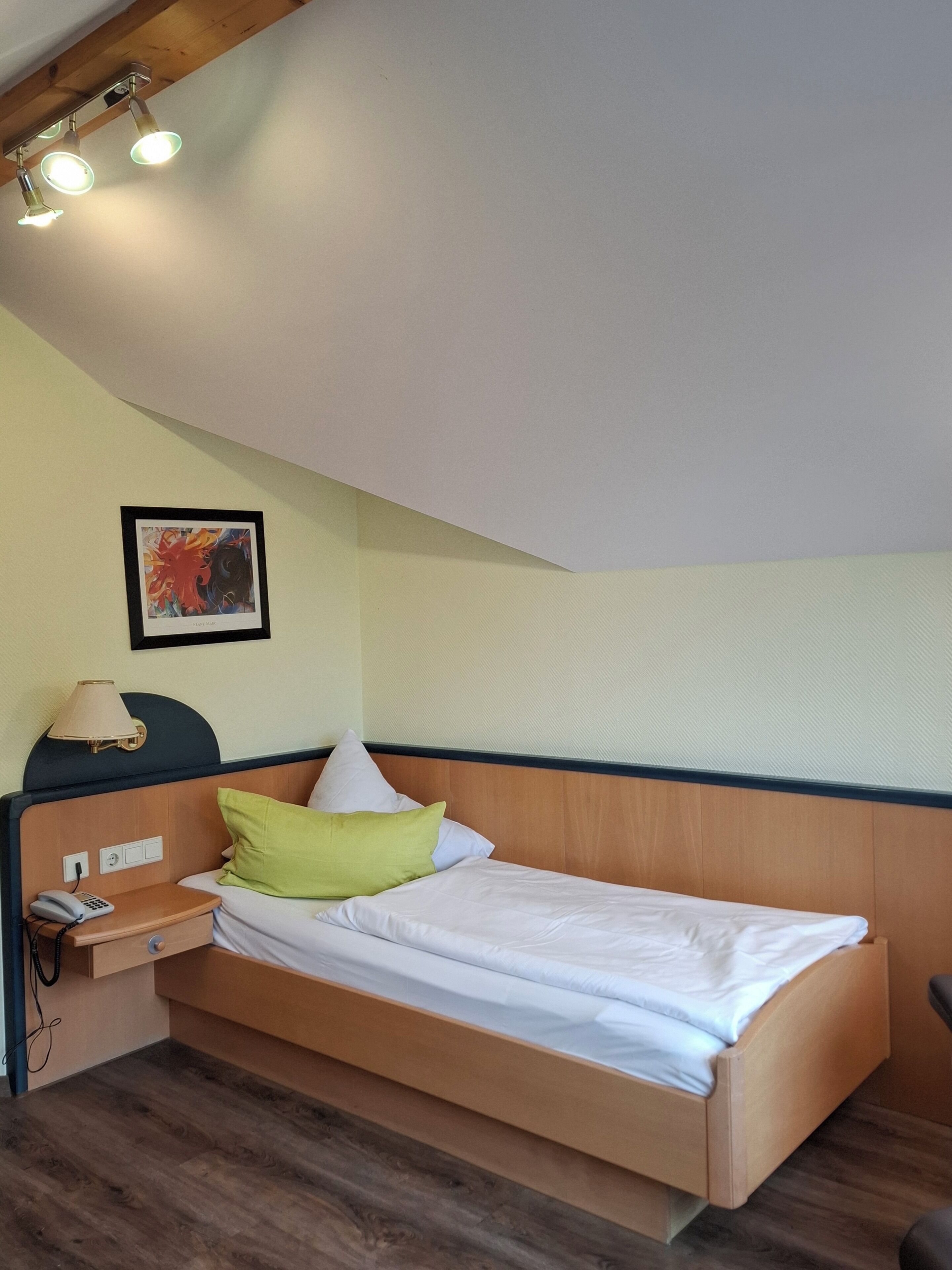 A single bed with a wooden headboard, a bedside table with a lamp, a phone, and a framed picture on the wall.