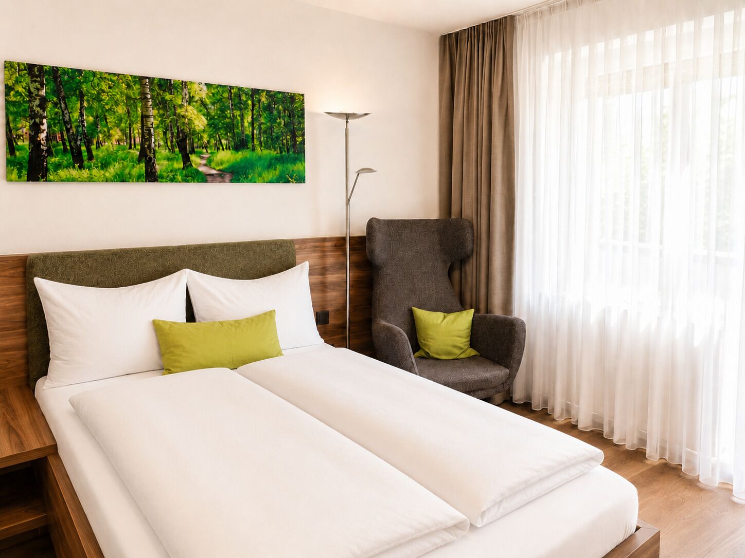 A hotel room with a bed, a chair, a painting of a forest, and sheer curtains.