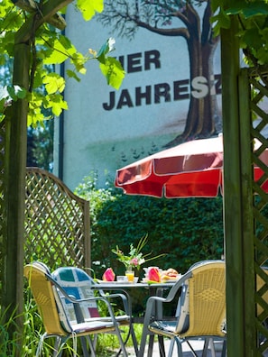 An outdoor seating area with wicker chairs and a table set for two, under a red umbrella. - Vitalhotel Garni Vier Jahreszeiten (Bad Fuessing)
