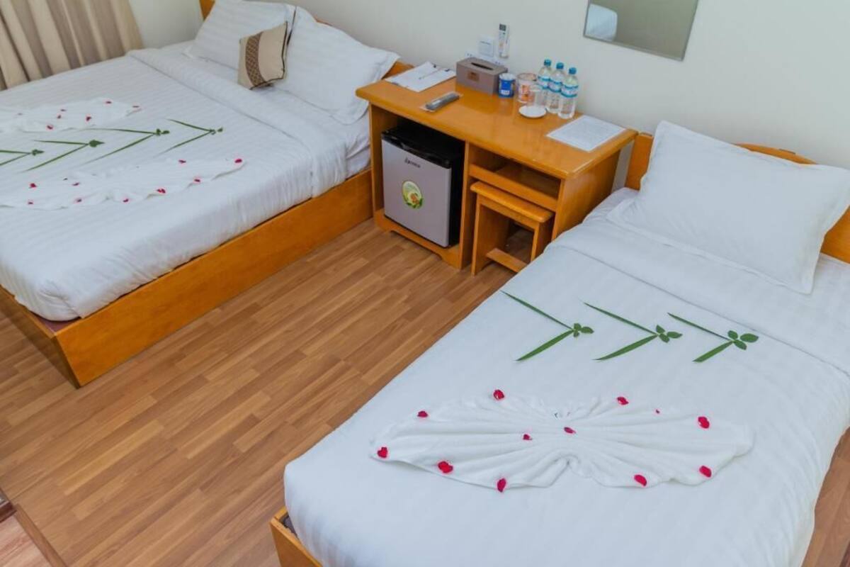 Une chambre d’hôtel avec deux lits, chacun recouvert d’une courtepointe blanche ornée de motifs floraux et de pétales de rose. Une table de chevet en bois se trouve entre les deux lits, sur laquelle on trouve un petit réfrigérateur, une table avec une télécommande, des bouteilles d’eau et un bloc-notes.