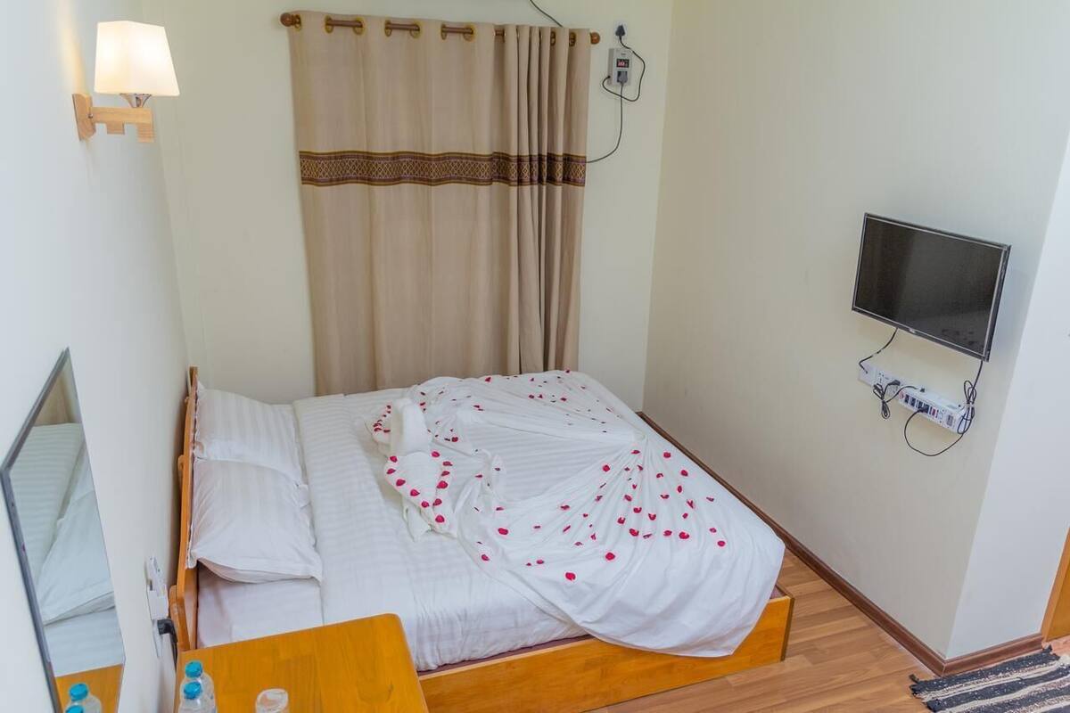 Un lit simple avec un couvre-lit blanc et des pétales de rose, une table de chevet avec une lampe, un téléviseur fixé au mur et une prise de courant.