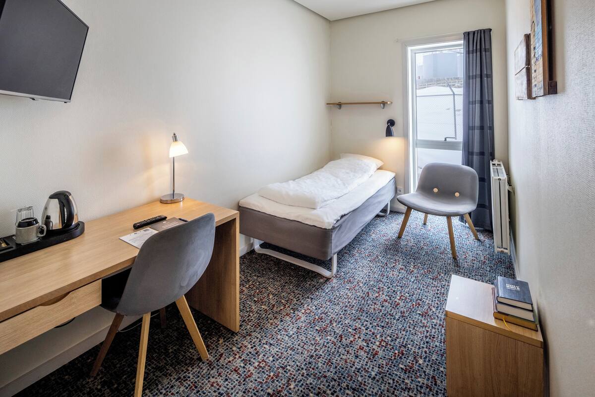 Une chambre d’hôtel de taille réduite, comprenant un lit simple, un bureau avec une chaise, une petite table avec une lampe et un radiateur.