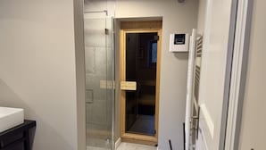 Een smalle gang met een glazen douchecabine, een houten deur naar een kamer en een witte radiator.