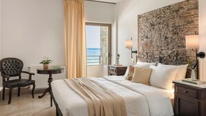 Habitación de hotel con una cama grande, una silla, una mesita con una planta, una mesita de noche con una lámpara y vista al mar a través de una ventana.