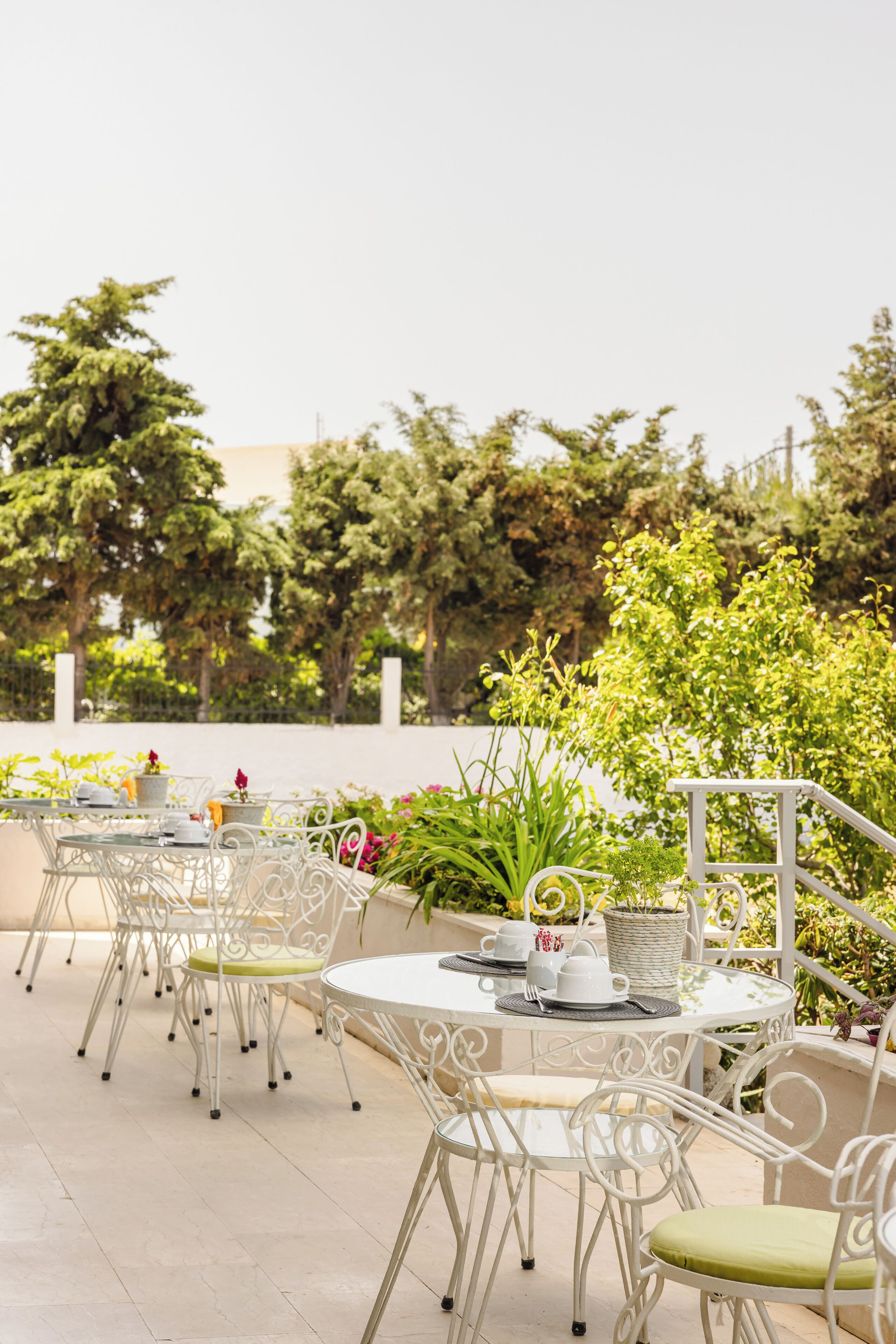 Outdoor seating with white metal chairs and tables, surrounded by greenery and trees.