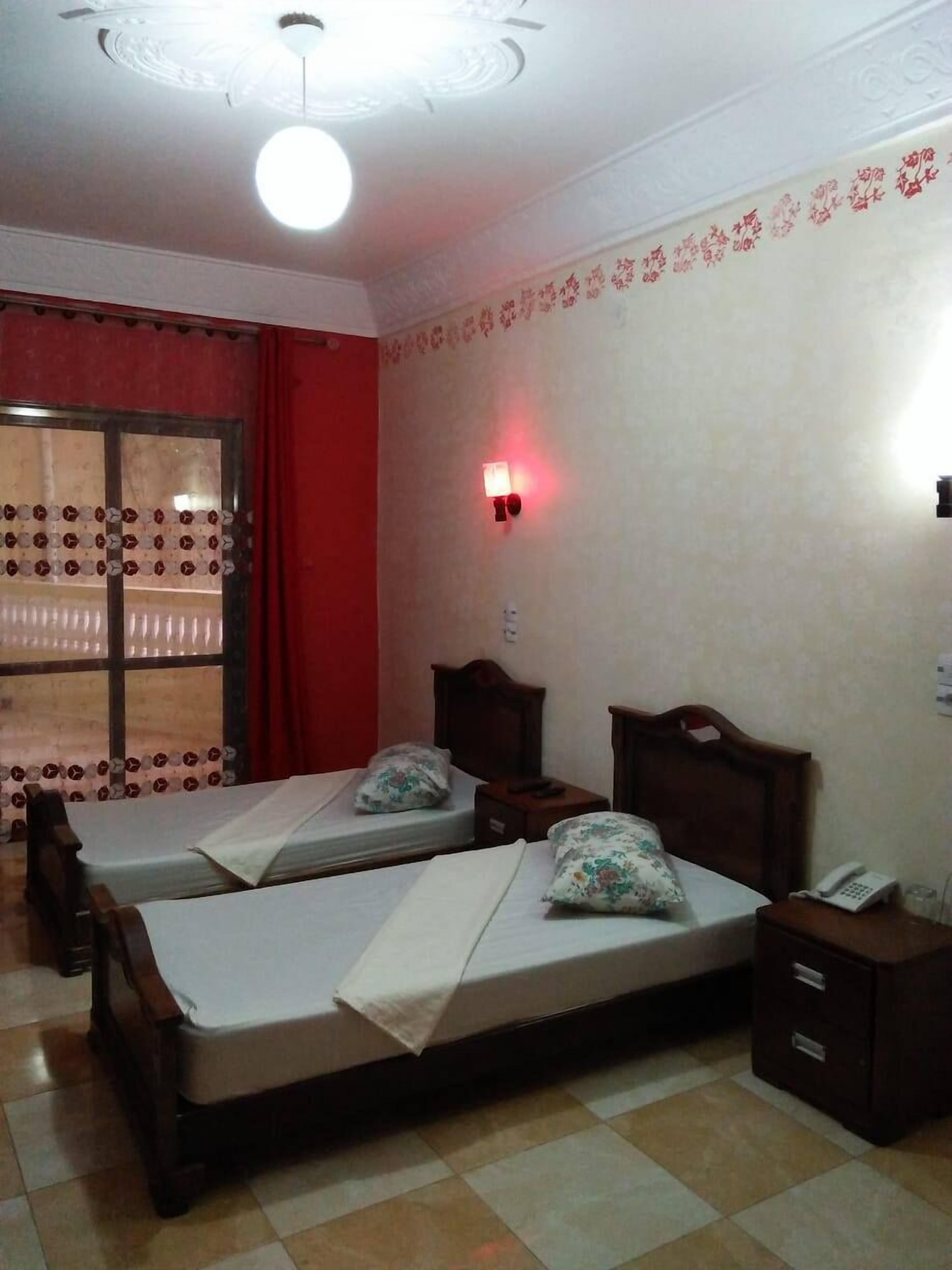 A room with two single beds, a red curtain, a wall-mounted light, and a window with patterned curtains.