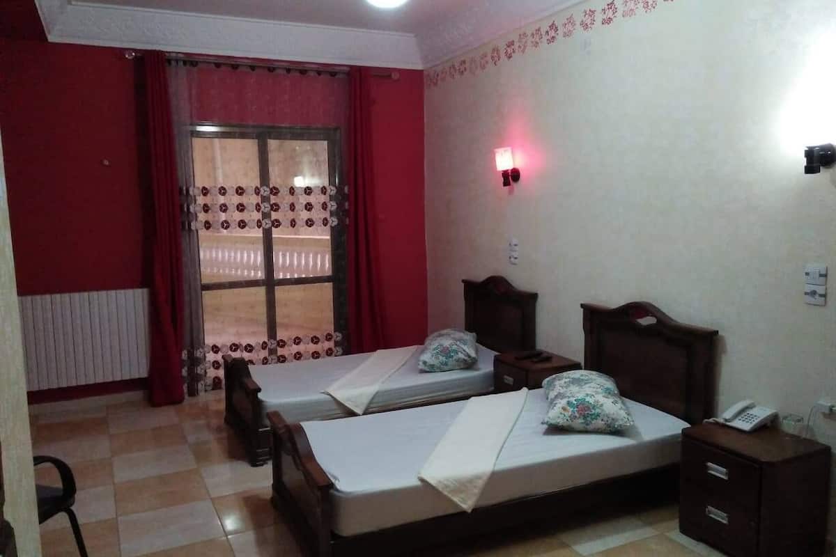 A room with two beds, a chair, a small table, a nightstand, a window with curtains, and a wall-mounted light.
