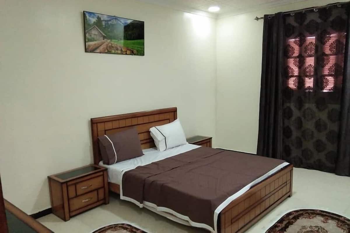 A bedroom with a wooden bed, two nightstands, a bedside table, a large painting on the wall, and a patterned curtain.
