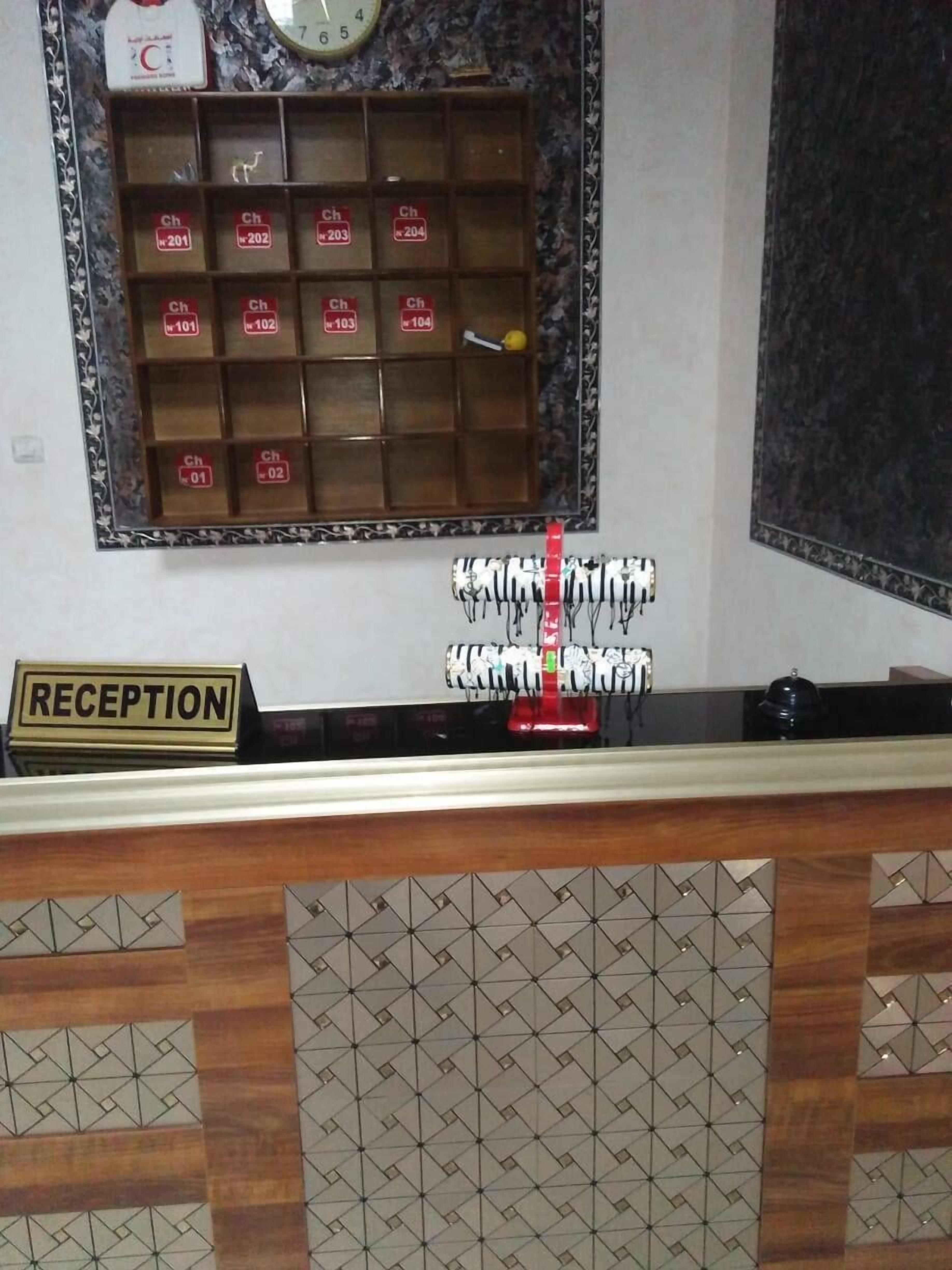 Zona de recepción con mostrador de madera, un letrero que dice 'RECEPTION', un reloj y una estantería con compartimentos numerados.