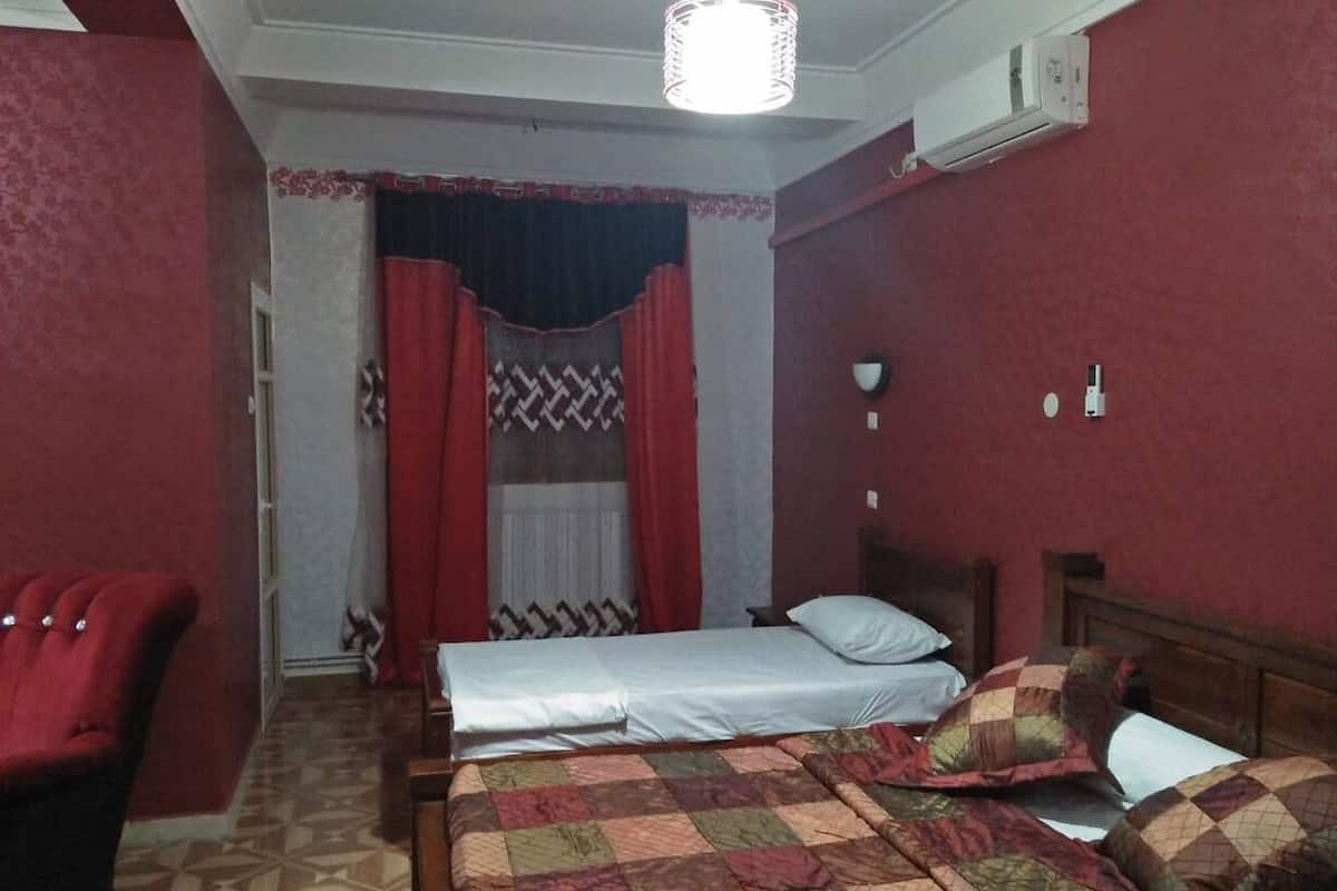 A bedroom with two beds, a red armchair, patterned curtains, and a ceiling light fixture.