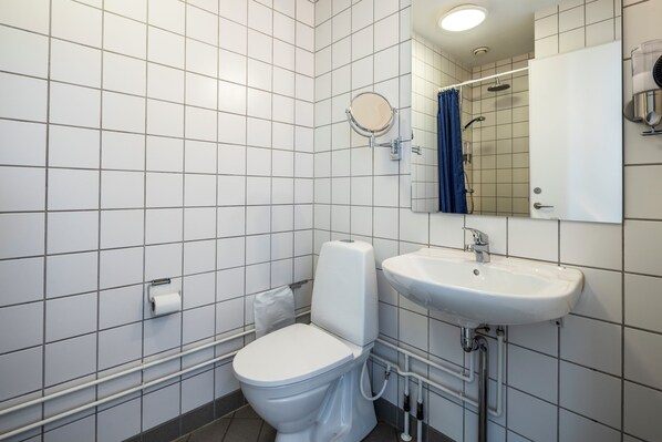 A modern bathroom with white tiles, a toilet, a sink, a mirror, and a shower area. - HOTEL SØMA Nuuk (Nuuk)