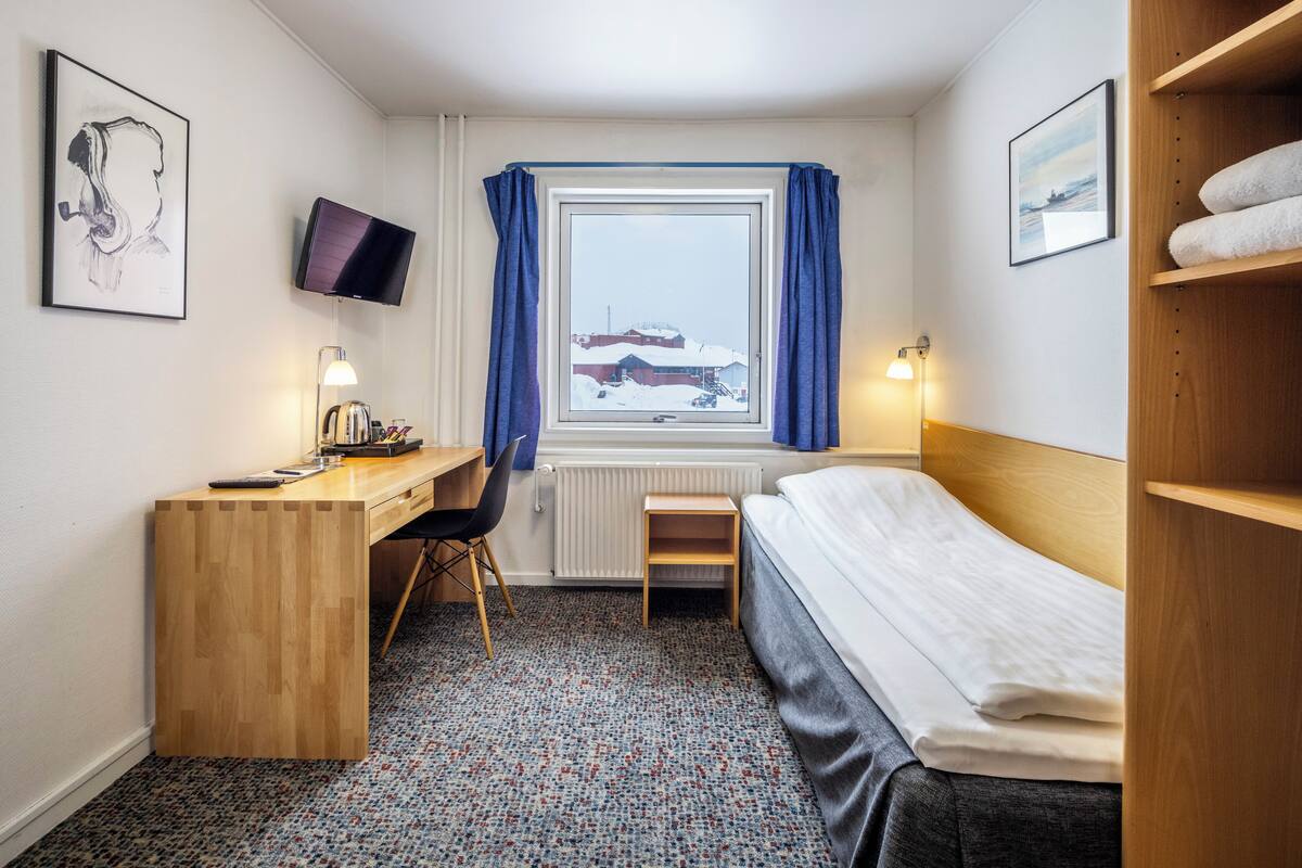 Ein kleines Hotelzimmer mit einem Holztisch, einem Einzelbett, einem Sessel, einem an der Wand montierten Fernseher und einem mit Vorhängen versehenen Fenster.