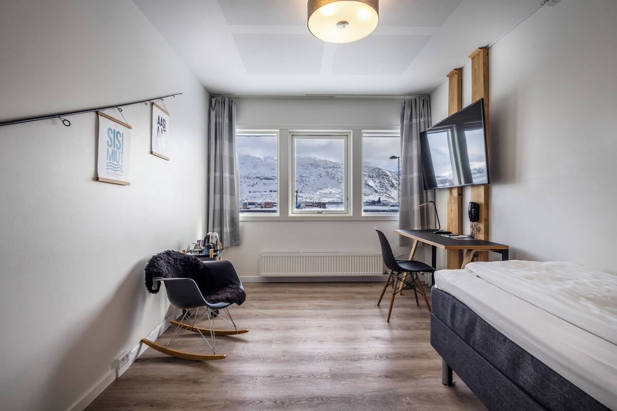 Ein modernes Hotelzimmer mit Bett, Schreibtisch, Stuhl und Blick auf schneebedeckte Berge durch große Fenster.