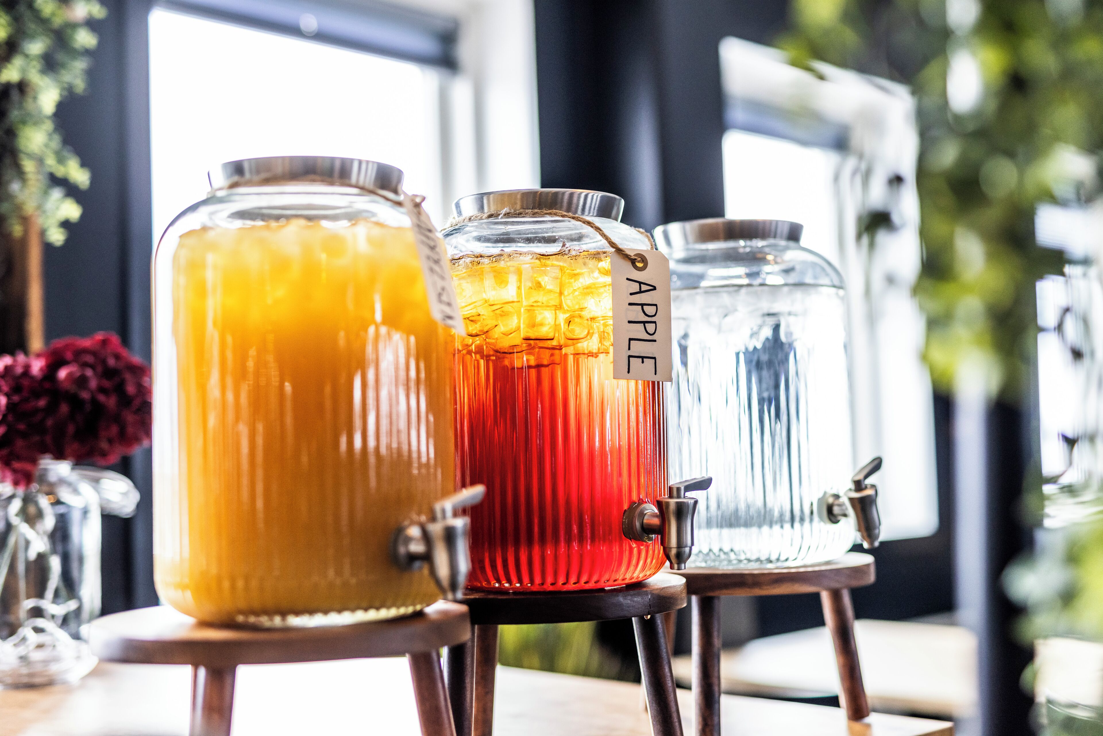 Three beverage dispensers with labels, one marked "APPLE," on a wooden table with a window in the background.
