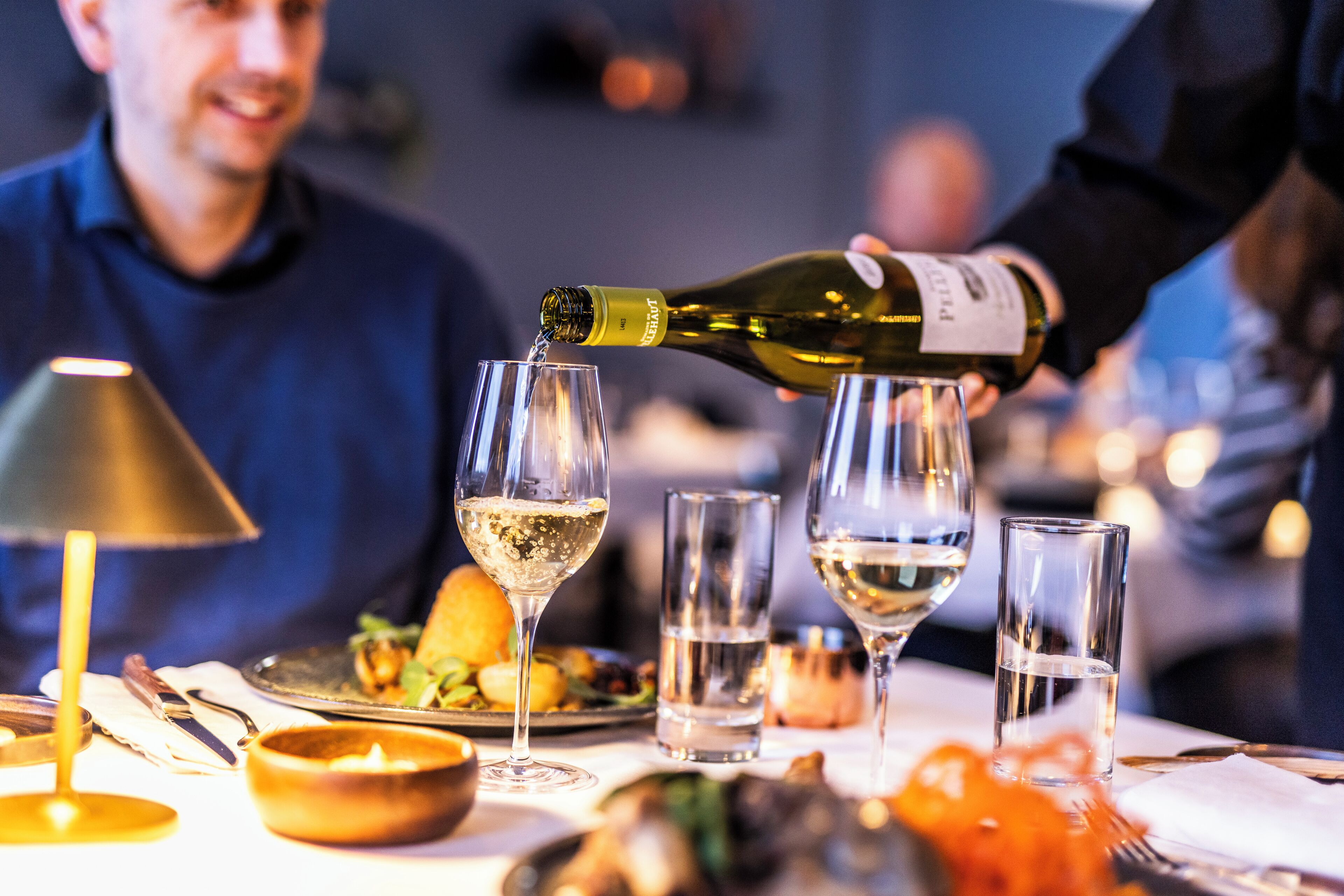 A dining table with a bottle of white wine being poured into glasses, a candle, and a plate of food.