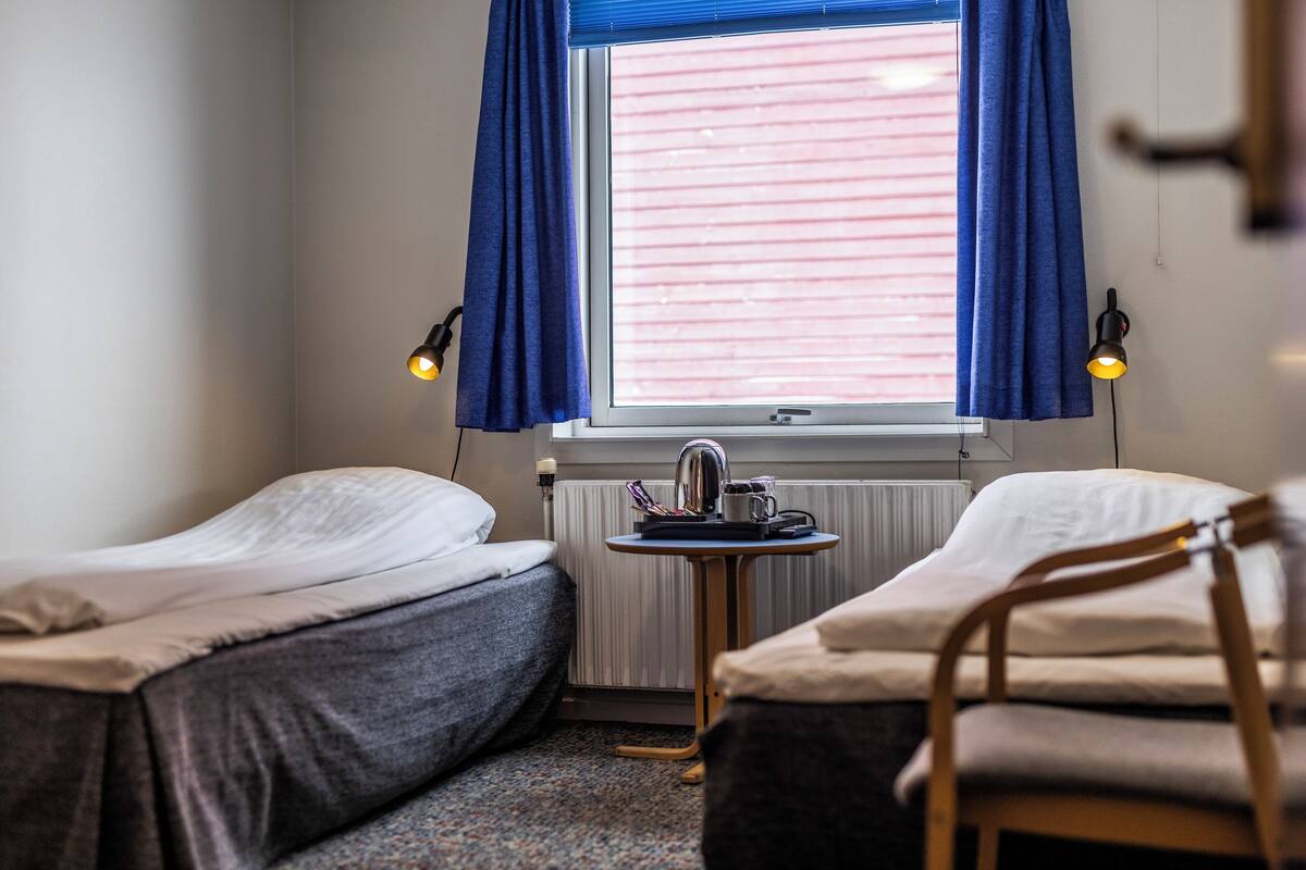 Ein Zimmer mit zwei Einzelbetten, einem kleinen Tisch mit Wasserkocher und einem Fenster mit Vorhängen.