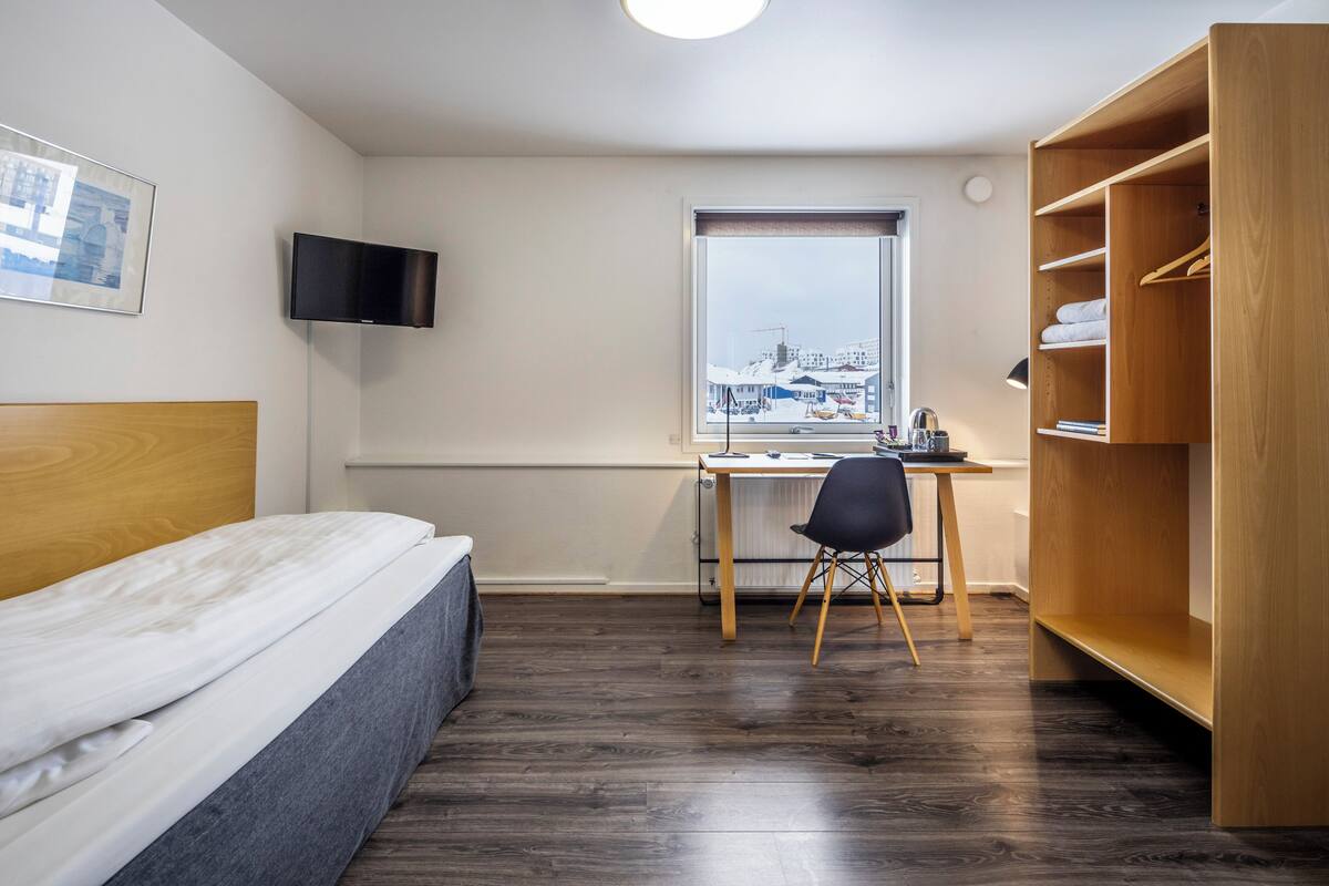 Ein Schlafzimmer mit Bett, Schreibtisch, Stuhl, Fernseher und Fenster mit Blick auf Gebäude.