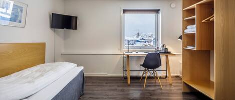Un dormitorio con cama, escritorio, silla, televisor y ventana con vista a edificios.