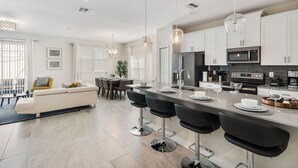 Eine moderne Küche mit weißen Schränken, einer zentralen Kochinsel mit Barhocker und einem Essbereich mit Sofa und Stühlen.