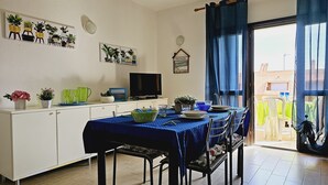 Un coin repas avec une table dressée pour quatre personnes, un téléviseur posé sur un meuble et diverses plantes en pot disposées sur le mur.