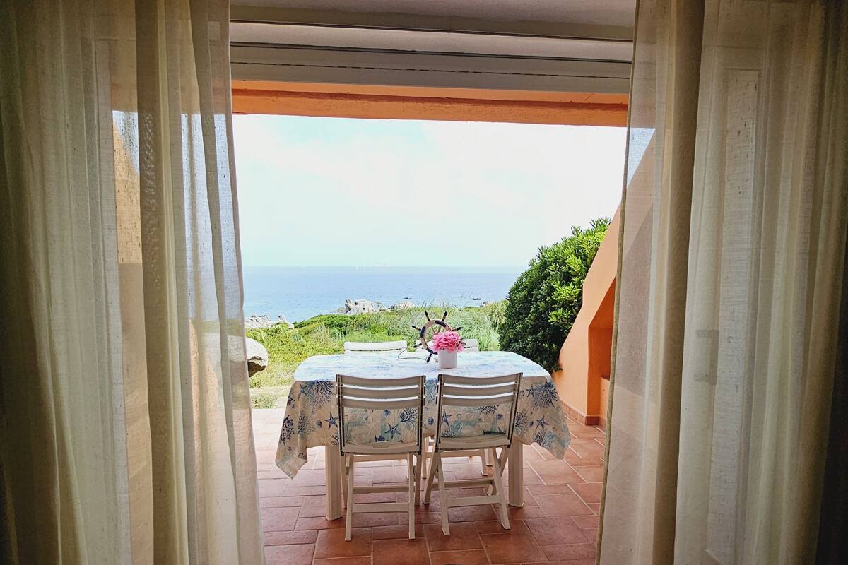 Eine Terrasse im Freien mit Tisch und Stühlen, Blick auf das Meer und einer mit Vorhängen versehenen Tür.