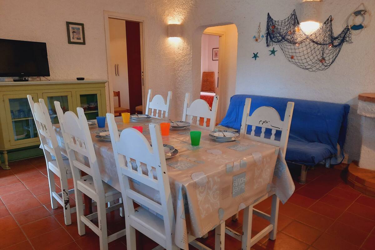 A dining area with a table set for four, surrounded by white chairs. There is a television on a cabinet to the left and a blue sofa to the right.