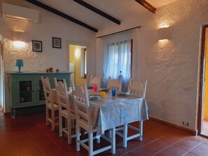 Uma área de refeições com uma mesa posta para quatro pessoas, rodeada por cadeiras brancas. Um armário verde com louças fica à esquerda.