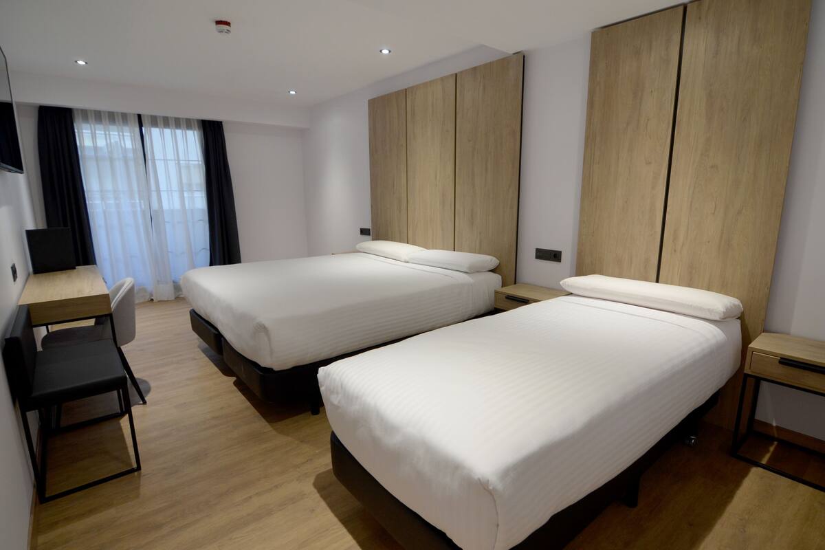 A modern hotel room with two single beds, a desk, a chair, and a wardrobe.