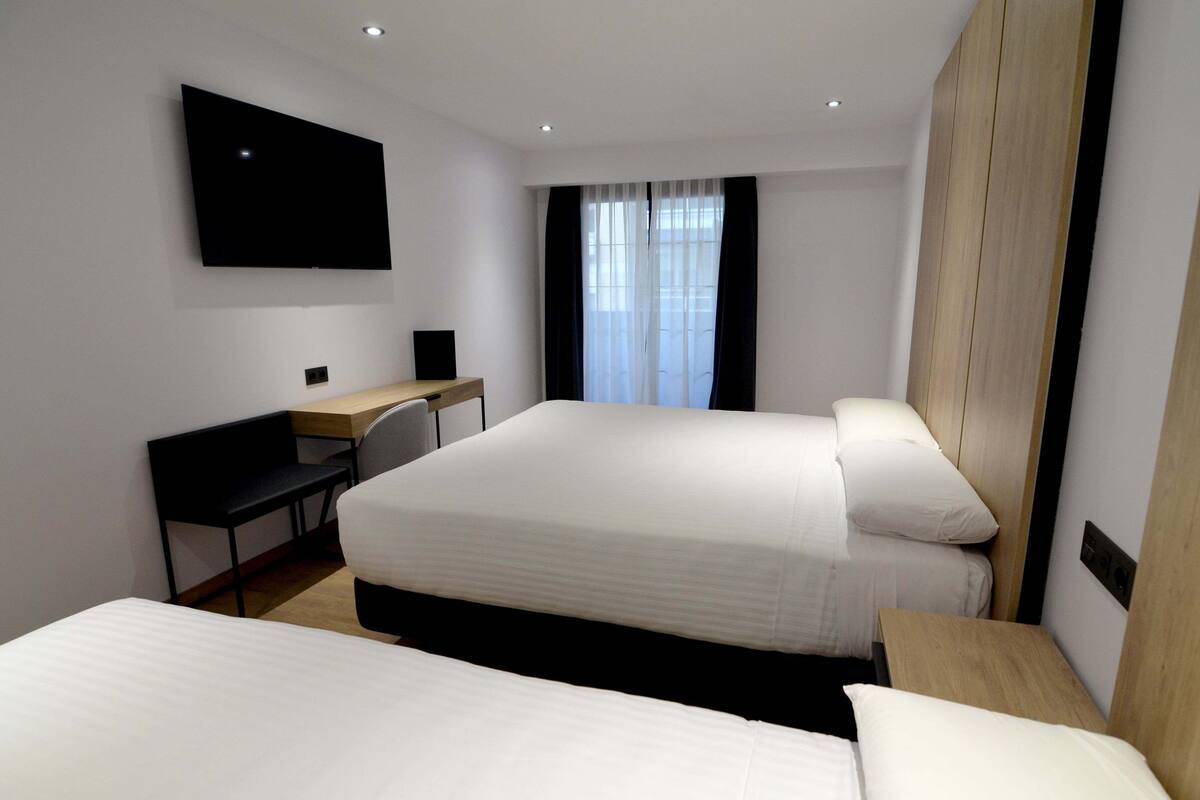 A hotel room with two beds, a desk, a flat-screen TV mounted on the wall, and a window with sheer curtains.