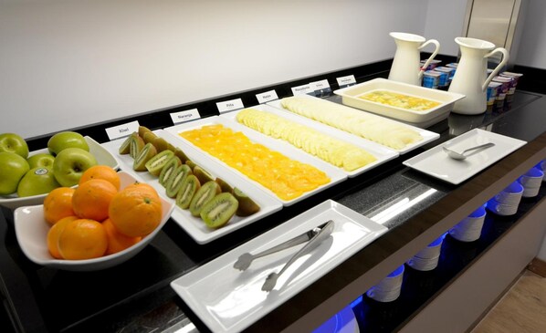 A buffet with labeled fruit and vegetable trays, a bowl of oranges, and a pitcher of water.
