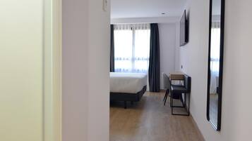 Habitación de hotel con cama, escritorio, silla, televisión y una ventana grande con cortinas.