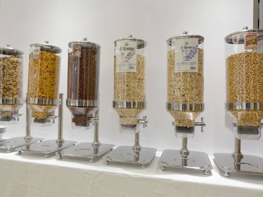 Seis dispensadores cilíndricos con tapas transparentes, cada uno conteniendo distintos tipos de cereales o granos, sobre una mesa blanca.
