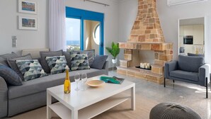 Ein modernes Wohnzimmer mit Kamin aus Stein, grauen Sofas, einem weißen Couchtisch und Blick durch eine blaue Tür.