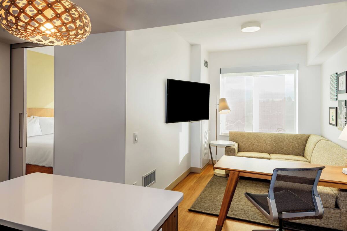 A modern hotel room with a flat-screen TV mounted on the wall, a sofa, a dining table, and a chair.