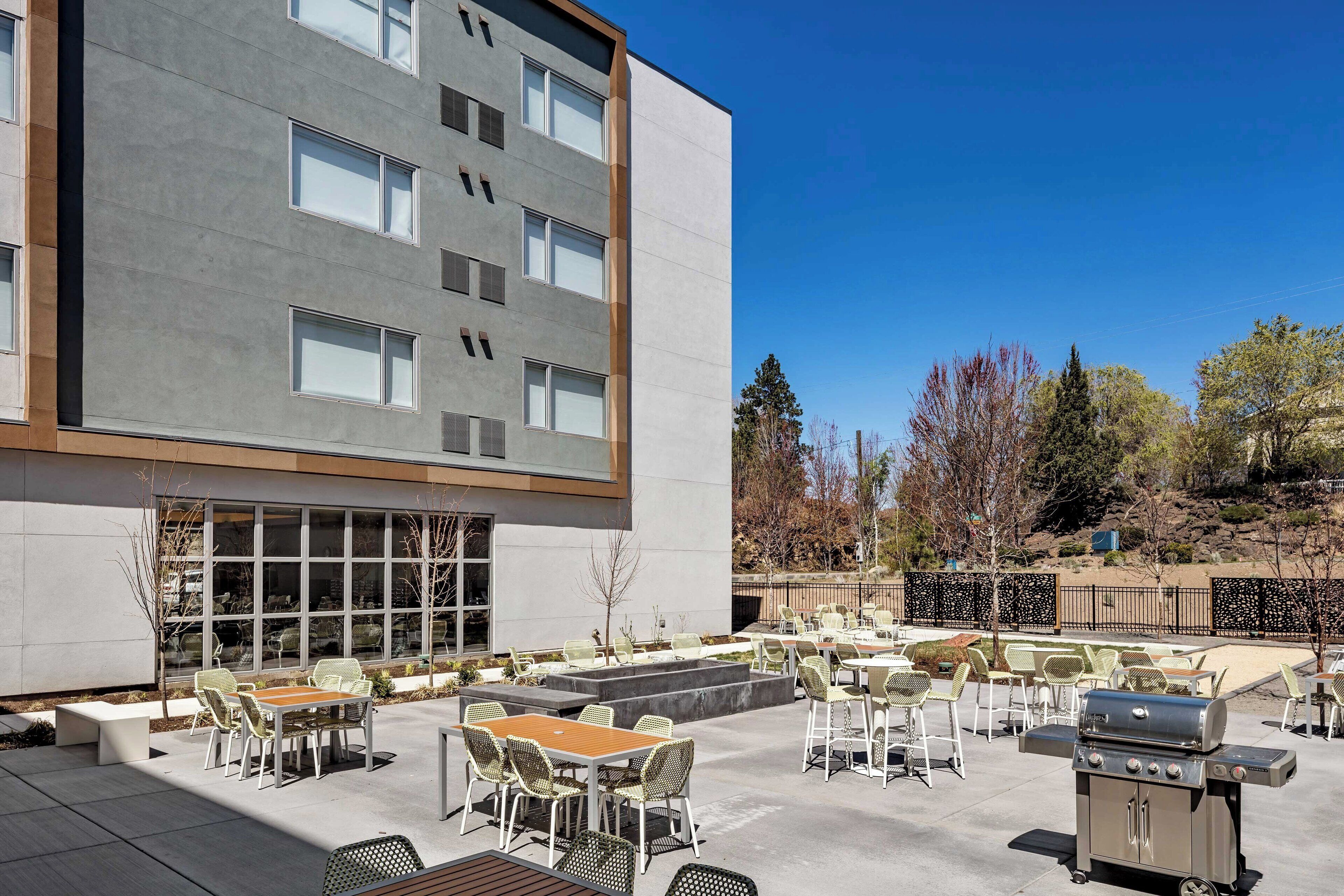 An outdoor patio area with multiple tables and chairs, a barbecue grill, and a building with large windows and a modern design.