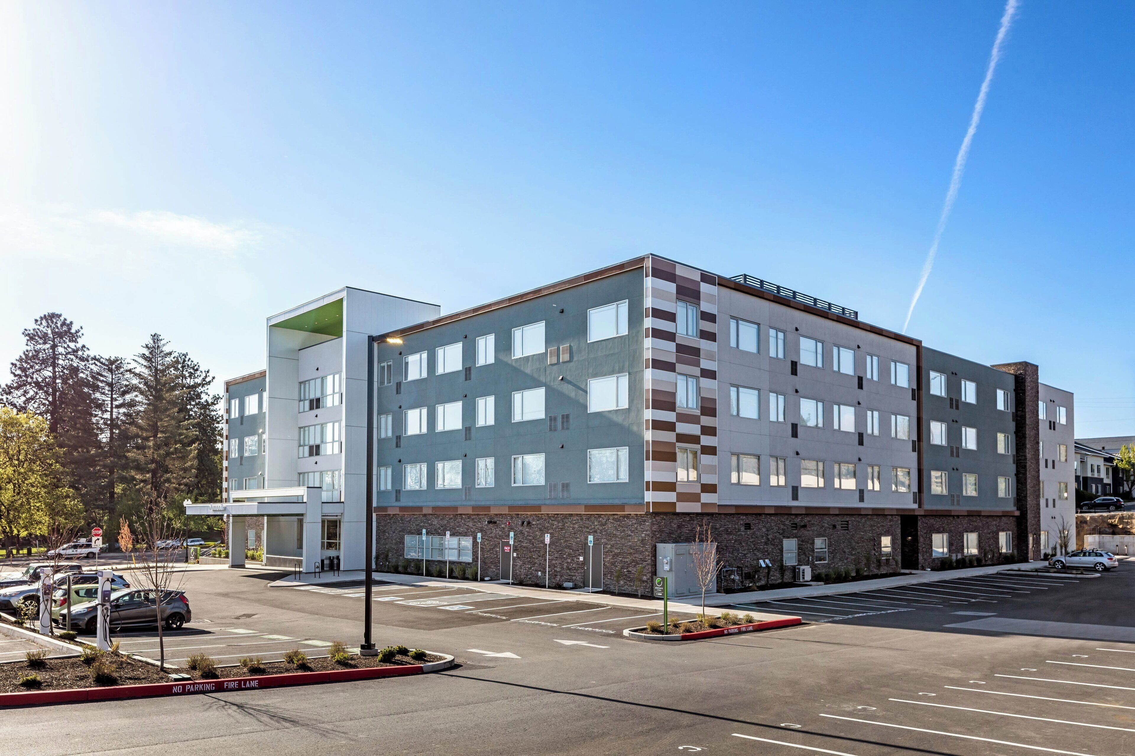 Ein mehrstöckiges Hotel mit modernem Design, inklusive Parkplatz mit eingezeichneten Stellplätzen und Feuerwehrzufahrt.