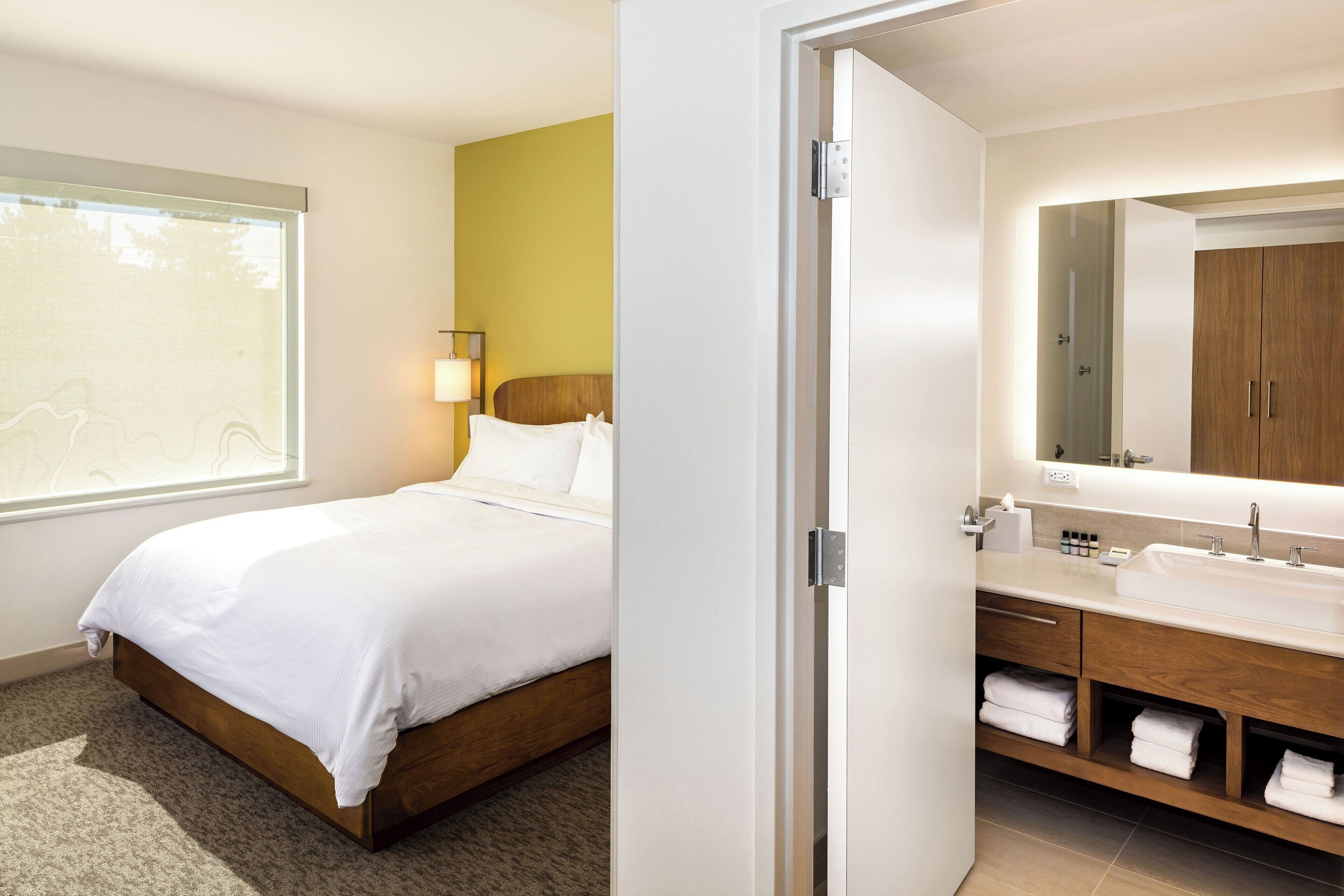 A modern hotel room with a bed, a bathroom with a sink and mirror, and a window with a view.