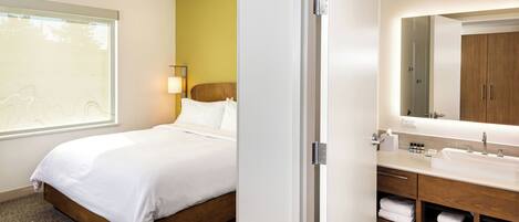 A modern hotel room with a bed, a bathroom with a sink and mirror, and a window with a view.