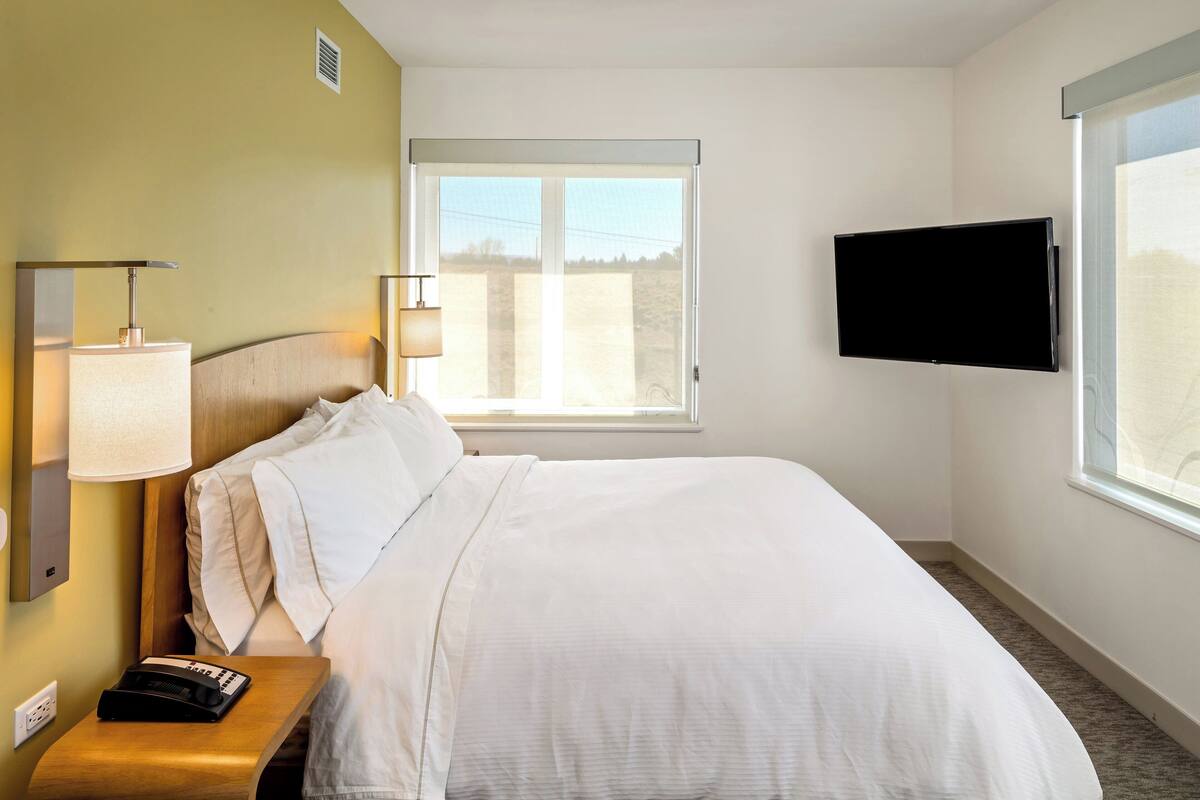 A hotel room with a bed, a bedside lamp, a telephone, a TV mounted on the wall, and a window with a view.