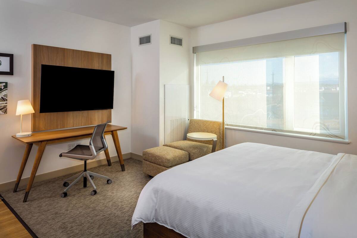 A hotel room with a bed, a desk with a chair, a TV mounted on the wall, a small round table with a lamp, and a window with a view.