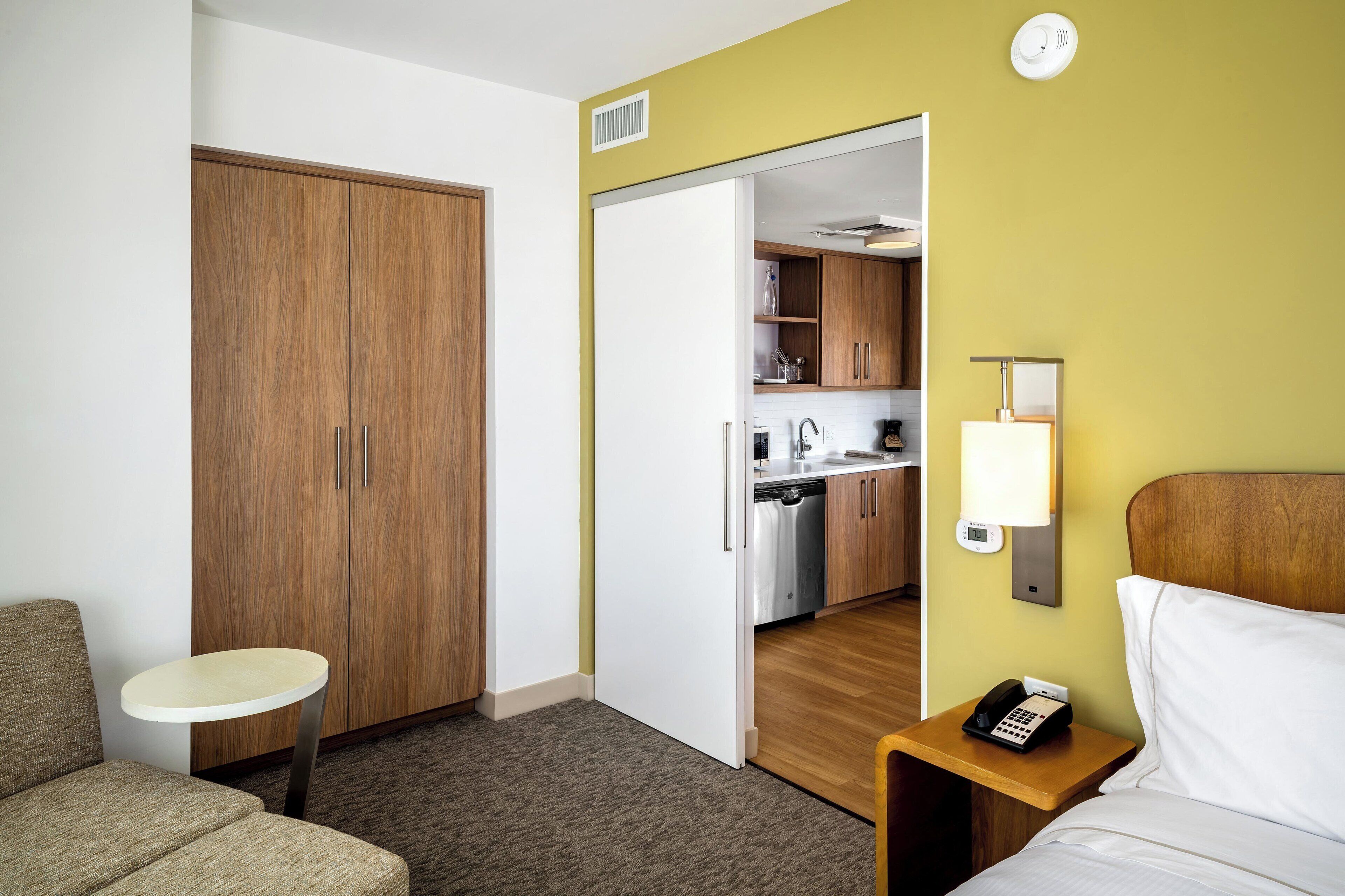 Une chambre d’hôtel comprenant un lit, une petite table, une armoire, un coin cuisine et une lampe de chevet.