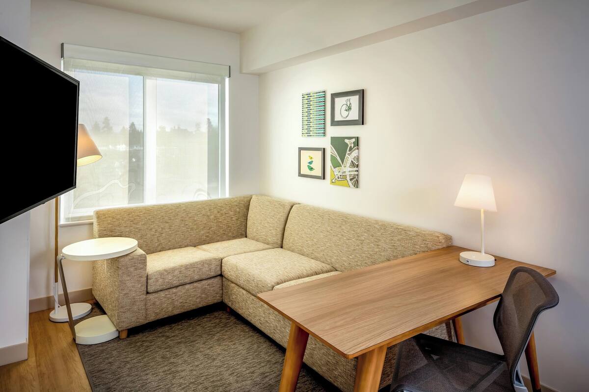 A modern living room with a beige sectional sofa, a wooden dining table, a small round side table, and a flat-screen TV mounted on the wall.
