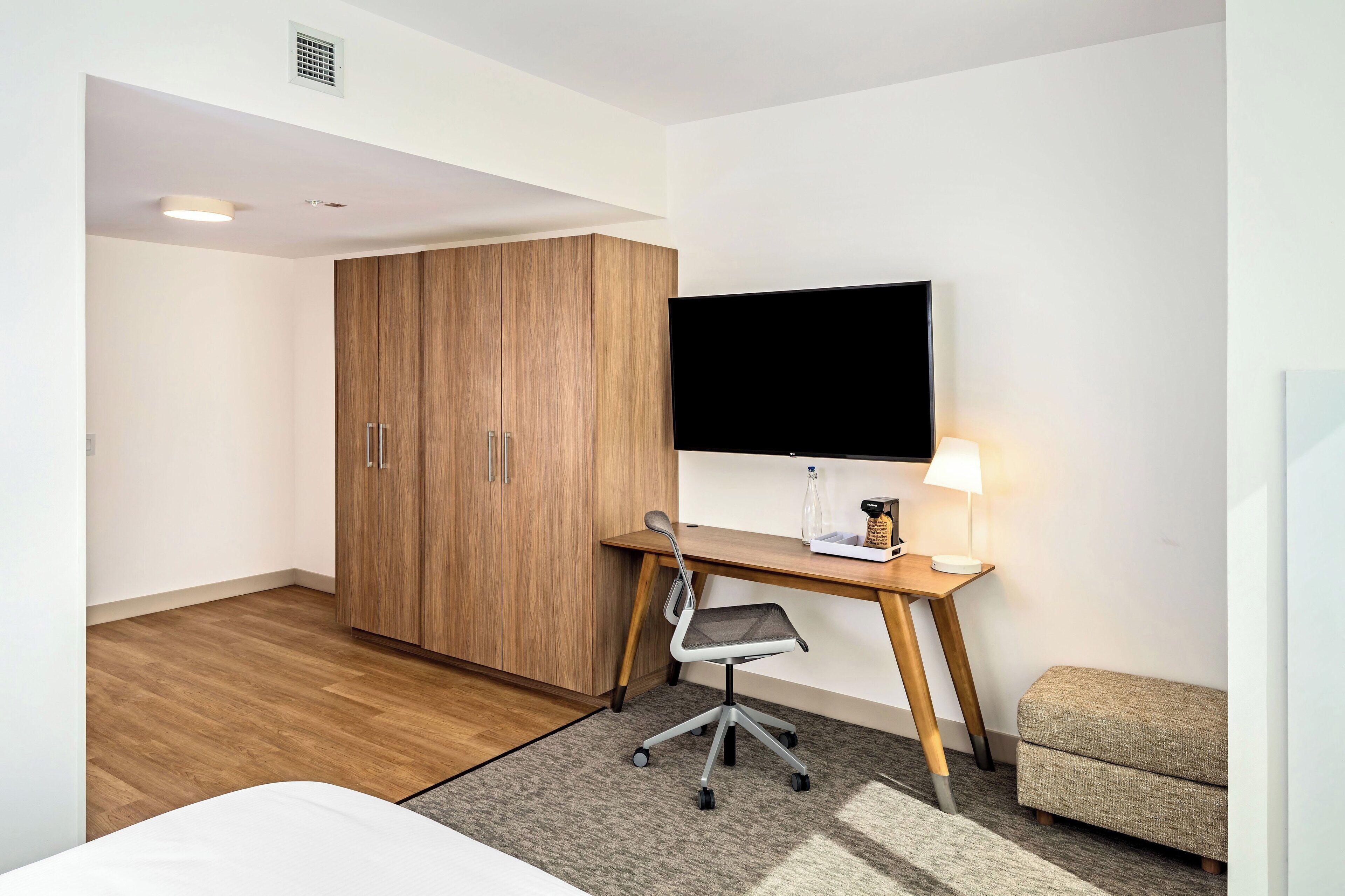 Een hotelkamer met een houten garderobe, een bureau met stoel, een flatscreen televisie, een nachtkastje met lamp en een kleine zithoek.