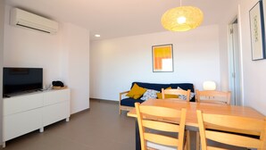 Un salon moderne avec un téléviseur posé sur un meuble blanc, un canapé bleu agrémenté de coussins jaunes, une table à manger en bois avec des chaises, et un luminaire suspendu.
