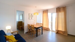 Un salon avec un canapé bleu, une table à manger en bois entourée de chaises, un luminaire suspendu et une vue sur l’extérieur grâce à de grandes fenêtres ornées de rideaux.